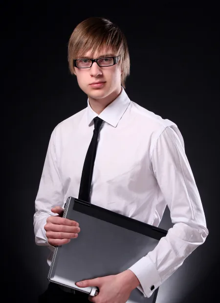 A man wearing glasses is holding a laptop, looking focused and engaged in his work.