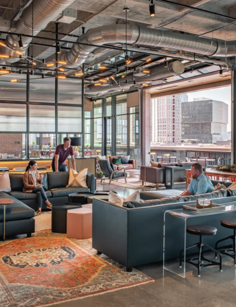 Modern office lounge with people conversing on dark sofas. Features a colorful rug, open ceiling with ducts, bright windows, and cityscape view, creating a relaxed, collaborative vibe